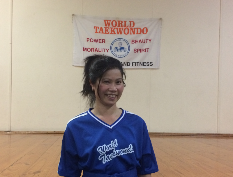 This photograph captures a smiling woman (My Tran) standing inside the training centre at Semaphore South Australia. She has medium-length dark hair tied back in a ponytail and is wearing a blue World Taekwondo T Shirt with white stripes on the sleeves. The T-Shirt has the words 