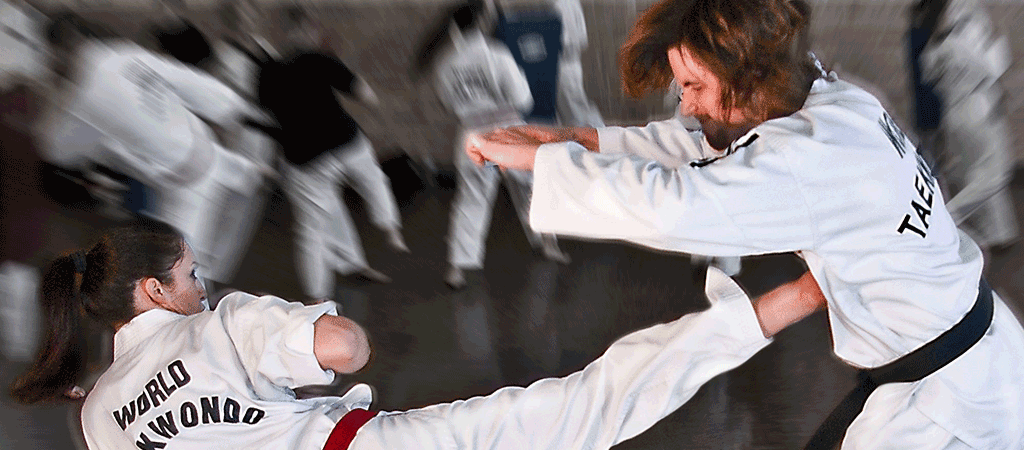 This is a dynamic photograph capturing a moment during a World Taekwondo training session. The central focus is on two individuals engaged in a sparring match. The person on the left, a young woman with dark hair tied back, is wearing a white Taekwondo uniform with the text WORLD TAEKWONDO in bold black letters on the back. She is executing a powerful side kick with her right leg extended, demonstrating high agility and strength. Her left arm is bent, providing balance. The person on the right, a man with brown hair, is also wearing a white Taekwondo uniform with black trim and the name WORLD TAEKWONOD in black letters on the back. Both participants are in mid-action, their expressions focused and determined. The background is slightly blurred, emphasizing the action in the foreground. The setting appears to be a training hall with a wooden floor and a few other individuals in white uniforms scattered around, engaged in various World Taekwondo movements. The overall atmosphere is one of intense focus and physical exertion, highlighting the discipline and athleticism involved in World Taekwopndo training.