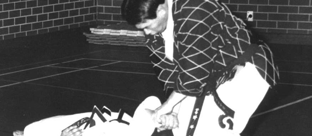 The image is a black-and-white photograph capturing a Hapkido training session with Grand Master Cha Sok Park. The central figure is Grand Master Park himself, wearing a traditional Hapkido uniform. The uniform is adorned with a black checkered pattern. He is in a kneeling position, with his hands gripping the arms of Wahid Halimee who is lying on the ground. 

The background features a brick wall, indicating an indoor setting. The floor is covered with a dark mat, and there are wooden floorboards visible on the right side of the image. The lighting is even, suggesting it is well-lit, typical of indoor training spaces. The overall atmosphere is focused and intense, reflecting the serious nature of this martial art. The textures of the uniforms, the mat, and the brick wall are all distinct, adding depth and realism to the photograph.