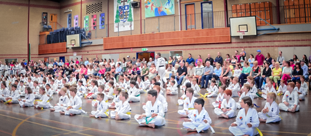 This photograph captures a lively indoor World Taekwondo event held in a large gymnasium. The gymnasium features a high ceiling with wooden beams and a large, colorful mural on one wall. The floor is polished hardwood with visible markings for various sports. 

In the foreground, numerous children and young adults are seated in rows on the floor, facing the front of the gym. They are dressed in white World Taekwondo uniforms with different colour belts, indicating varying levels of experience. Some students hold yellow belts, which are worn around the waist. The students are seated cross-legged, with some deep in concentration or meditation.

The background is filled with spectators, including adults and children, seated on benches. The audience is dressed in casual attire, with a mix of bright colors like pink, blue, and yellow. The spectators are engaged, watching the students attentively.

The atmosphere is vibrant and focused, with the students' white uniforms standing out against the gym's neutral tones. The photograph captures the sense of community and dedication to world taekwondo training.
