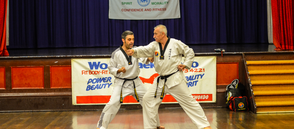 World Taekwondo demonstration taking place on a wooden stage with a dark, polished floor. Two men are engaged in a dynamic pose, both dressed in white Taekwondo uniforms with black belts. The man on the left has short, dark hair and is striking a powerful stance with his left foot forward, while the man on the right, with short, grey hair, is executing a similar stance but with his right foot forward. Both men are wearing black belts with white stripes, indicating their proficiency in martial arts. In the background, a large banner hangs from the stage, featuring the text World Taekwondo Fitness in blue and red letters against a white background. The stage backdrop is dark blue with a few decorative panels. To the right of the stage, there is a black bag with a red logo and a black backpack resting on the floor. The stage is surrounded by deep red curtains that extend to the floor, adding a touch of elegance to the setting. The overall atmosphere suggests a formal, organized event. The lighting is bright, highlighting the participants and the stage area.