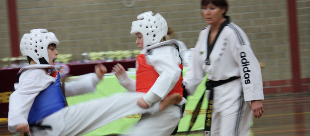 This photograph captures a dynamic moment in a World Taekwondo competition. In the foreground, two young students, both wearing white uniforms with black belts and protective headgear, are engaged in a competition bout. The student on the left is wearing a blue protective vest, while the one on the right has a red protective vest. Both students are in mid-action, with the student on the left executing a kick, their leg extended high in the air, and the other student slightly off-balance, preparing to block the kick.

In the background, Instrructor Margaret Rogers in a white World Taekwondo uniform with black stripes, also wearing a black belt, is refereeing. Behind her, a table covered with a green cloth is visible, with several trophies or awards arranged neatly on top, indicating a competitive or achievement-focused environment.

The setting is an indoor gymnasium with a high ceiling and beige brick walls. The floor is a polished wood finish. The overall atmosphere is focused and energetic, reflecting the intensity and discipline of World Taekwondo training.