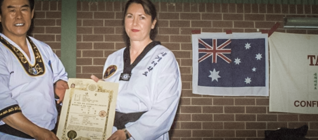 This photograph captures a moment in Australia. The main subjects are two people standing side by side. On the left is grand master Cha Sok Park with a medium build and short, dark hair, wearing a traditional Taekwondo uniform adorned with black trim and a distinctive sash. He is smiling warmly. On the right is Master Carol Halimee with a medium build and shoulder-length brown hair, also dressed in World Taekwondo uniform with black trim and a black belt. She is holding a certificate in her right hand, presented to her by the grand master. The certificate is framed in gold and features a seal and official text in Korean.

The background consists of a brick wall with a large Australian flag mounted on it. The flag is positioned to the right side of the image and is hanging on a piece of cloth that is pinned to the wall. The flag is predominantly white with a blue union jack in the upper left corner and the Australian coat of arms in the lower right. To the right of the flag, there is a partially visible red banner.