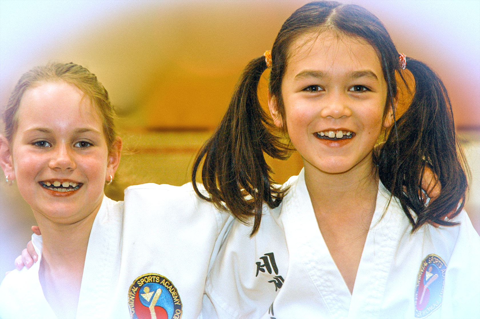 This is a photograph featuring two young girls smiling broadly and standing close together. Both girls are wearing traditional World Taekwondo uniforms, which are adorned with a blue and red emblem on the left breast pocket. The girl on the left has her light brown hair tied back in a simple ponytail, while the girl on the right has her hair styled in two pigtails, secured with red hair ties. The girl on the left has a slight, joyful smile, while the girl on the right is laughing heartily, showing her teeth. Their expressions suggest a sense of camaraderie and enjoyment. The background is blurred, with soft, warm lighting that creates a welcoming atmosphere. The overall mood of the image is cheerful and energetic, capturing a moment of joy and friendship between the two girls. The photograph has a soft, slightly vintage filter that adds a nostalgic and warm tone to the image.